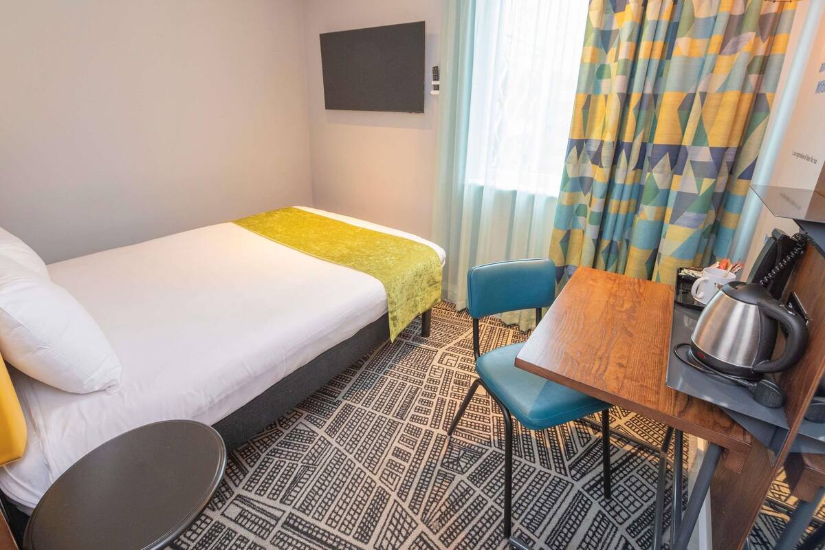 A hotel room with a bed, a desk with a chair, a kettle, and a patterned floor.