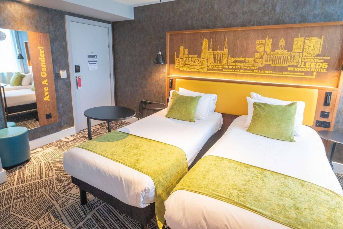 A hotel room with two beds, a small table, a chair, and a cityscape wall art.