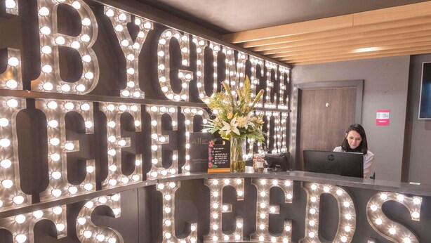 A reception desk with illuminated letters spelling 'BY YOUR SIDE IS LEEDS' and a receptionist behind the counter.
