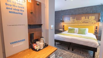 A hotel room with a wooden desk, a bed with a cityscape headboard, and a tray with tea and snacks.