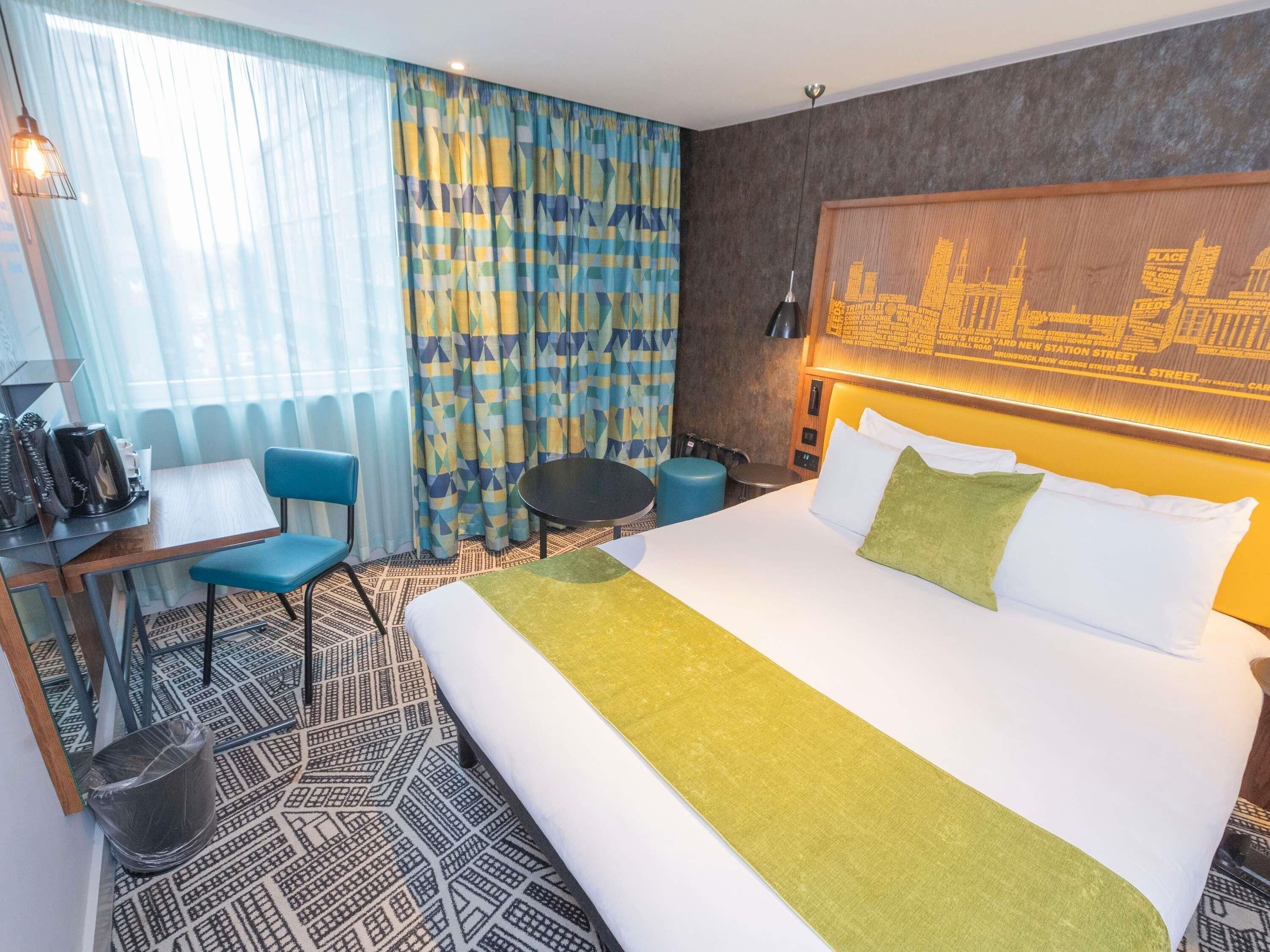 A hotel room with a bed, a desk with a chair, a small table, and a trash bin.
