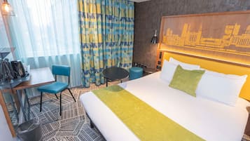A hotel room with a bed, a desk with a chair, a small table, and a trash bin.