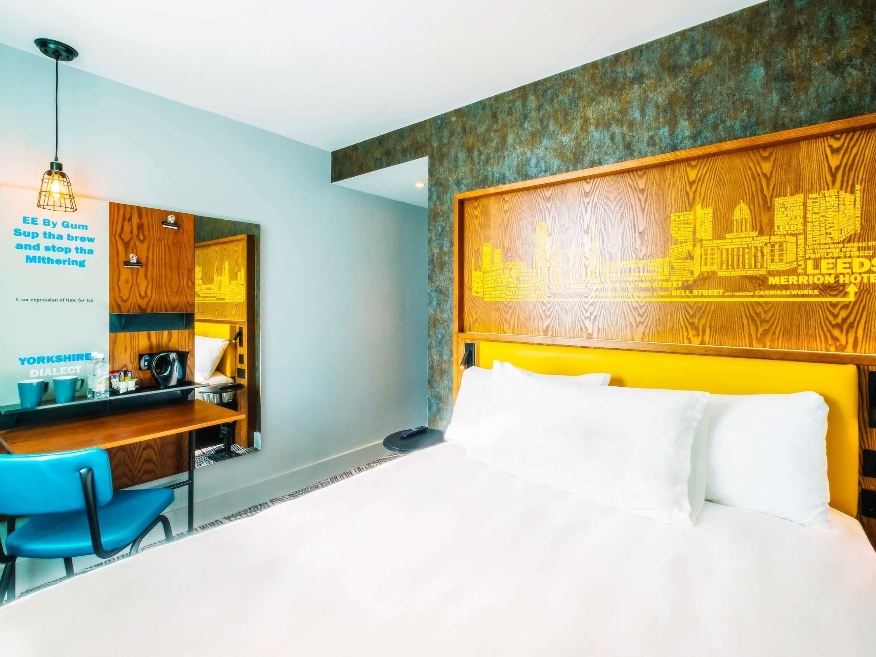 A hotel room with a bed, a desk with a chair, a mirror, and a cityscape mural on the wall.