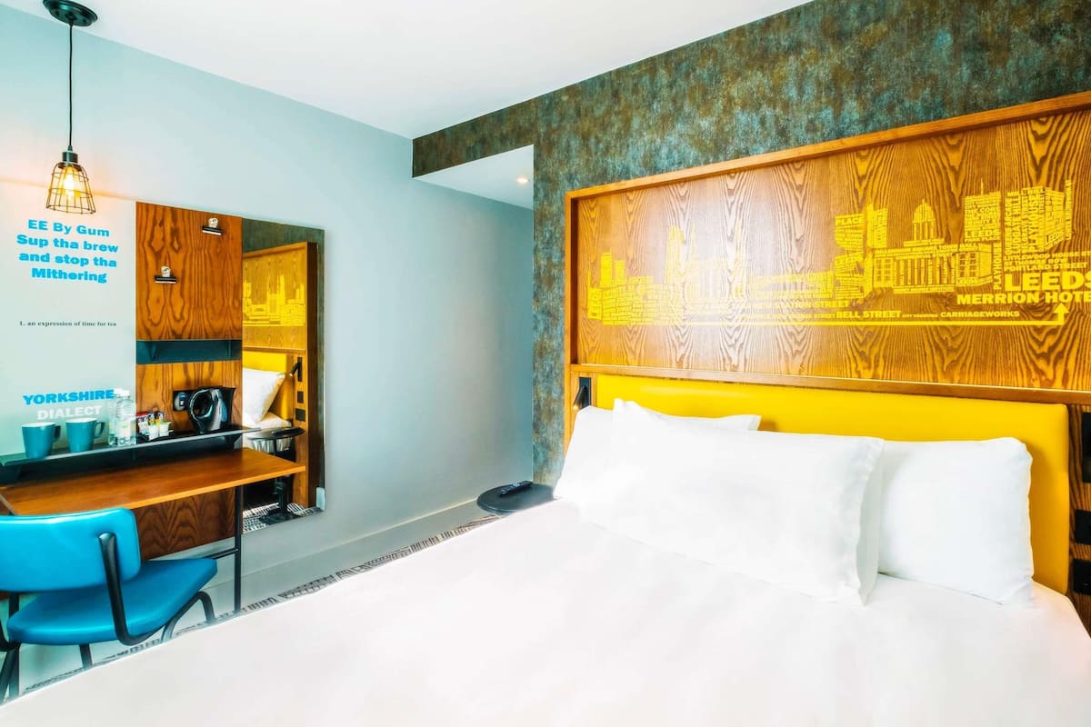 A hotel room with a bed, a desk with a chair, a mirror, and a cityscape artwork on the wall.