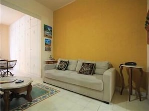 Ein Wohnzimmer mit einem beigefarbenen Sofa, einem kleinen runden Tisch und einem Couchtisch. An der Wand hängt ein Gemälde und es gibt ein Fenster mit Jalousien.