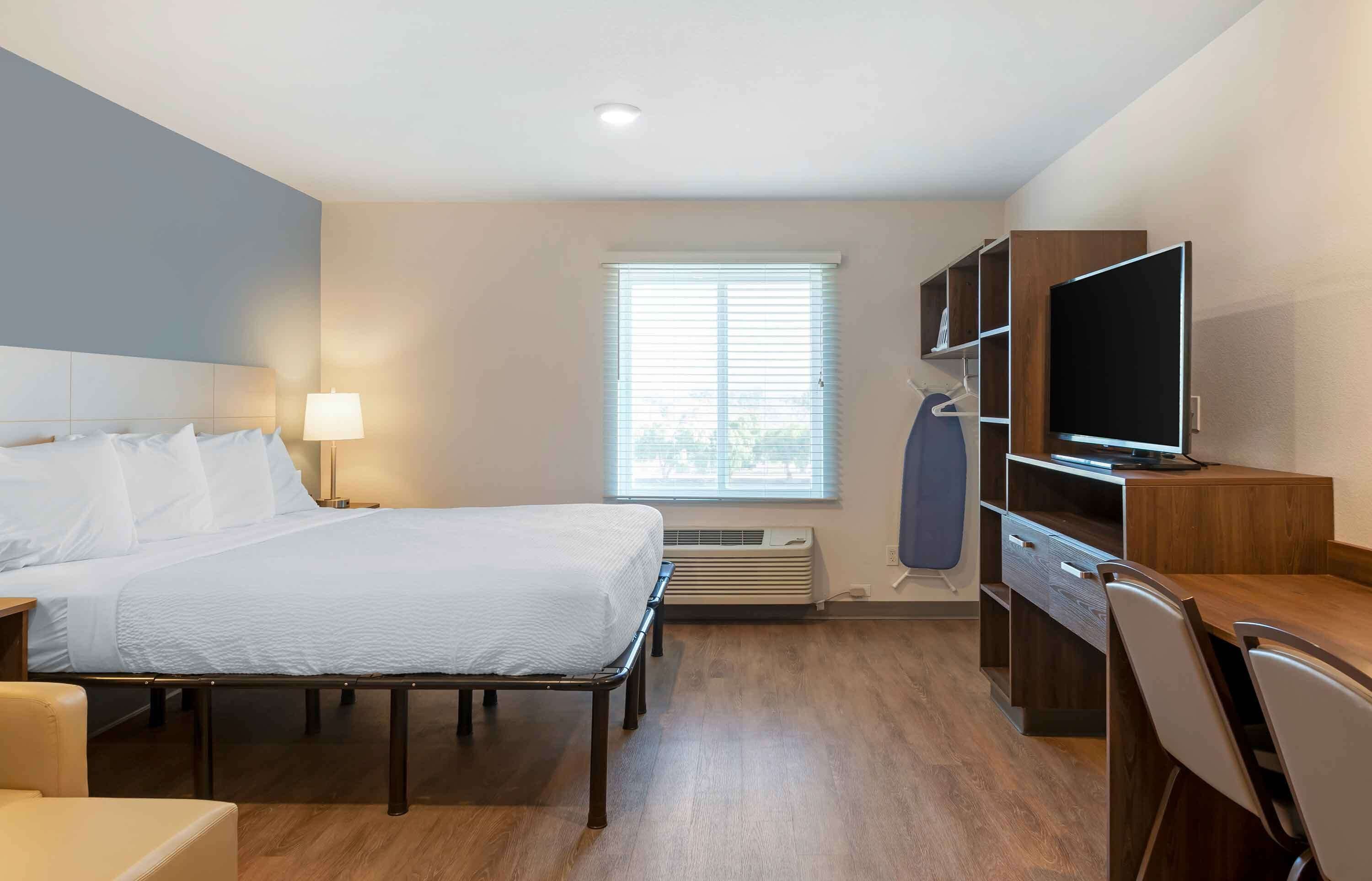 Habitación de hotel con cama, mesita de noche, lámpara, televisión, escritorio, silla y ventana con persianas.