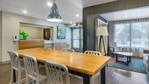 Una habitación de hotel con una mesa de comedor de madera, sillas blancas, una lámpara colgante moderna, un sofá, una lámpara de pie y un amplio ventanal con cortinas.