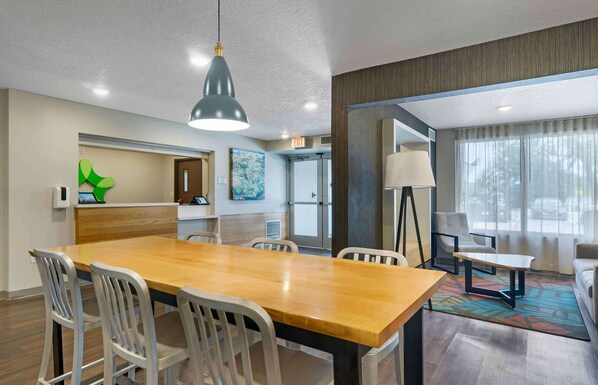 Una habitación de hotel con una mesa de comedor de madera, sillas blancas, una lámpara colgante moderna, un sofá, una lámpara de pie y un amplio ventanal con cortinas.