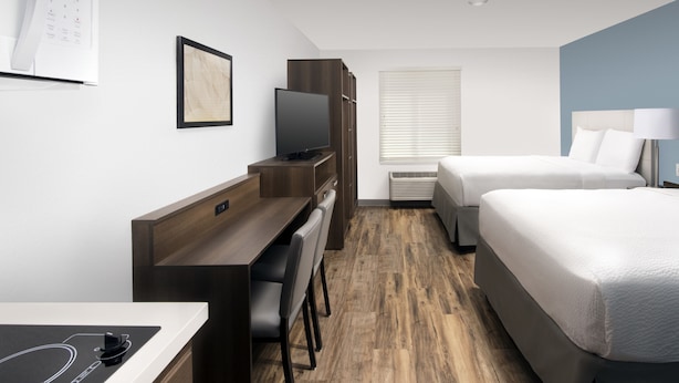 Una habitación de hotel moderna con un escritorio de madera, un televisor de pantalla plana, una cama con ropa blanca y una ventana con persianas.