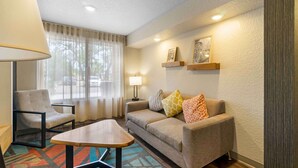 Una habitación de hotel acogedora con sofá, sillón y una pequeña mesa. Hay una ventana con cortinas, una lámpara y estantes en la pared con fotografías.