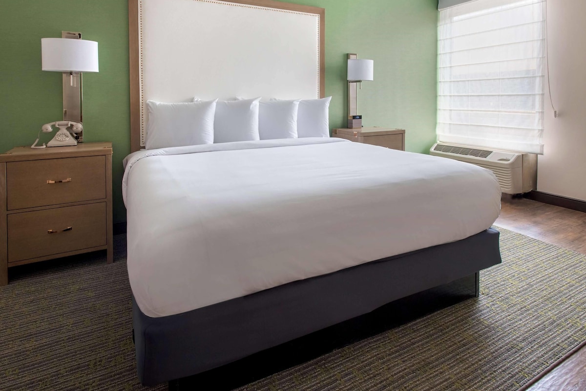 A hotel room with a large bed, two bedside tables, a headboard, and a window with blinds.
