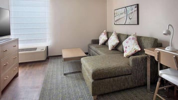 A compact hotel room with a sofa, a desk with a chair, a television, and a window with blinds.