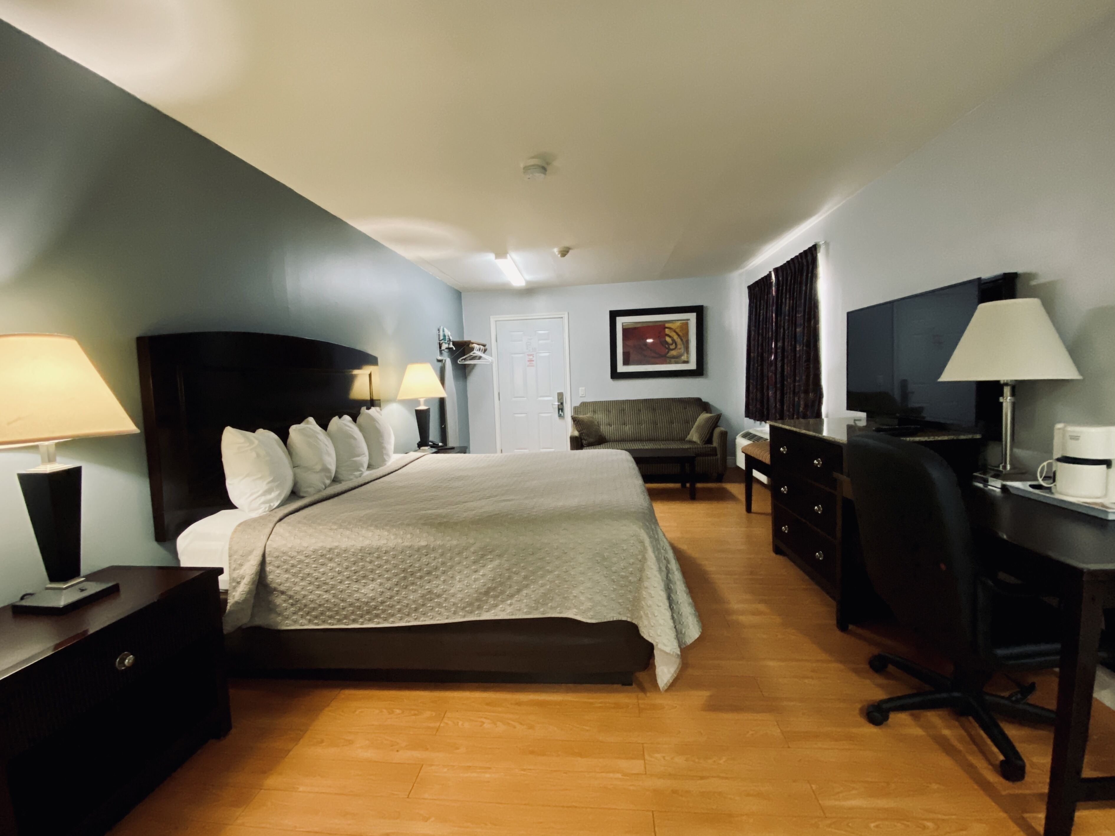 Une chambre d’hôtel avec un lit, un bureau, une chaise, une commode, une lampe, une machine à café et un tableau au mur.