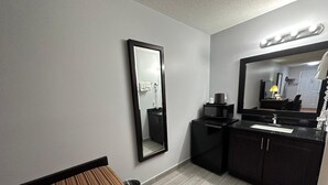 A compact bathroom with a large mirror, a sink, a microwave, and a bench.