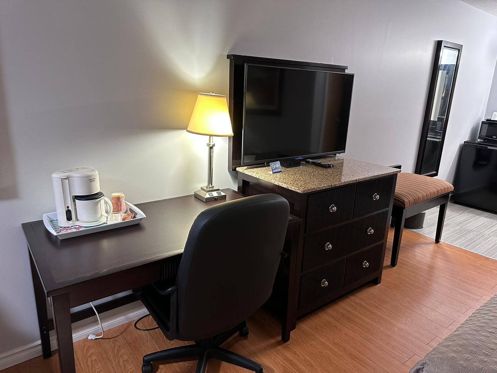 A hotel room with a desk, a chair, a television, a coffee maker, and a lamp.