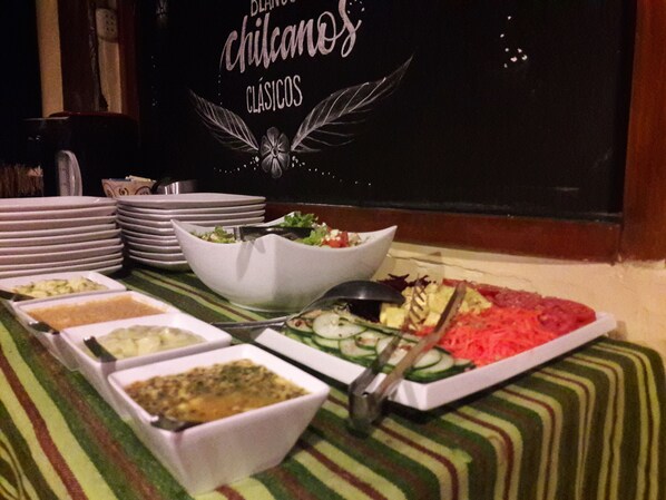 A buffet table with various dishes and bowls of food, a stack of plates, and a sign with the text "Blanco Chicanos Classics".