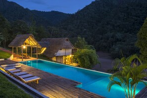Uma área à beira da piscina com espreguiçadeiras e um deck de madeira, cercada por vegetação exuberante e montanhas ao fundo.