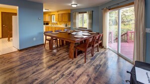 A dining area with a wooden table set for six, a kitchen in the background, and a large window with curtains.