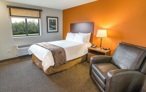 A hotel room with a bed, a nightstand, a lamp, a chair, and a window with a view of trees. - My Place Hotel - Boise/Meridian, ID (Meridian)
