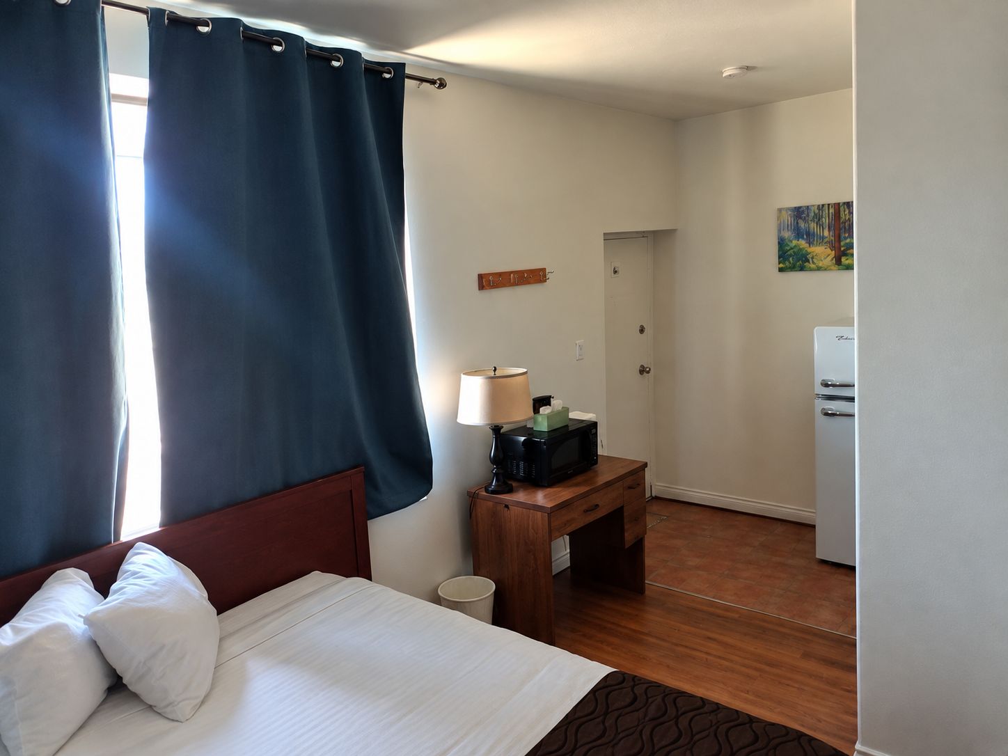 Quarto de hotel com cama, escrivaninha, luminária, micro-ondas e geladeira.
