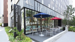 Een modern hotel met een glazen gevel, een terras met tafels en stoelen en twee parasols voor de schaduw.