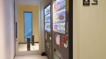 A hallway with vending machines on the right, a door at the end, and a blue-tinted window on the left.