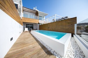 Una piscina moderna al aire libre con cubierta de madera y guijarros blancos, rodeada por una valla de madera y un muro blanco.