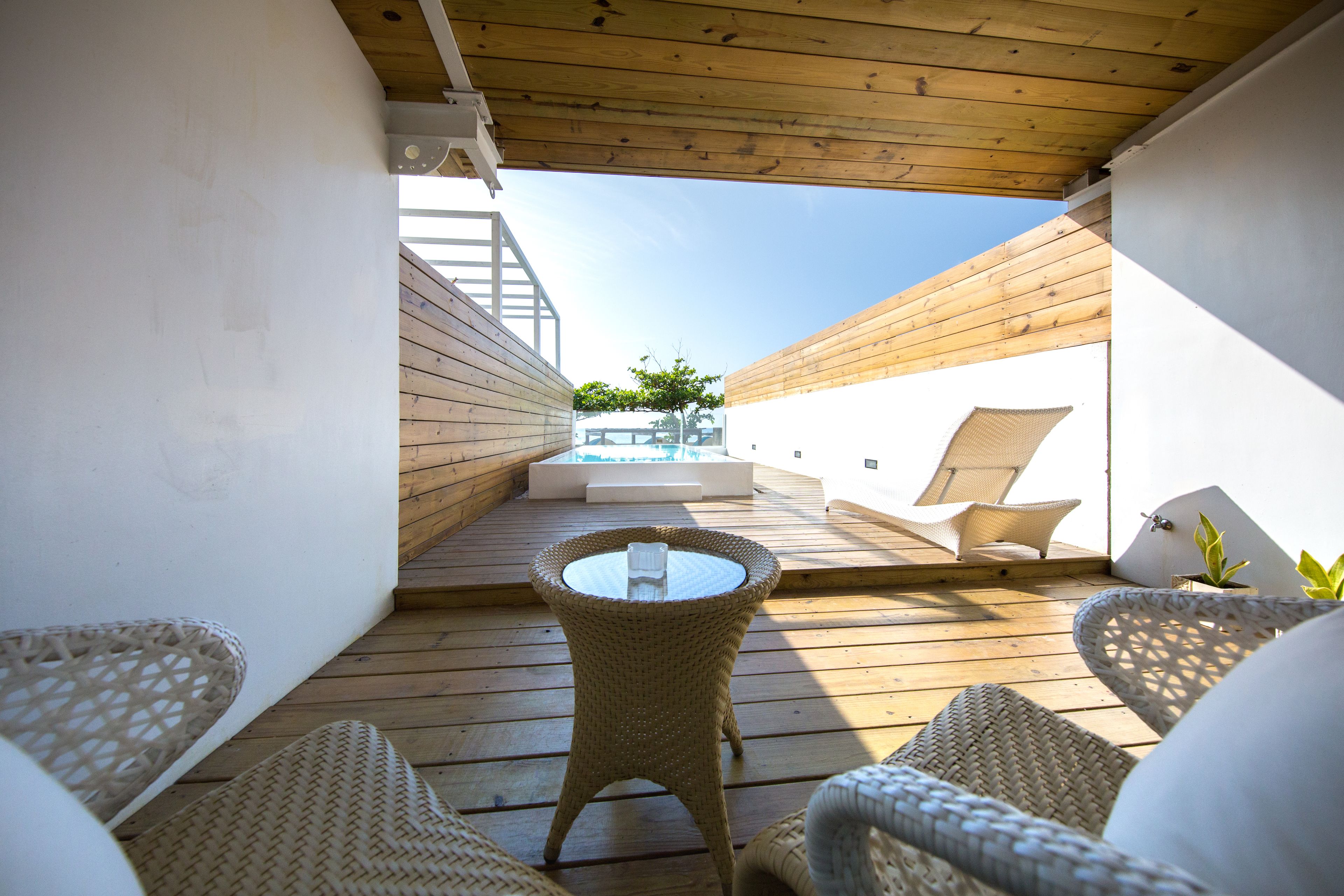 Une terrasse en bois, agrémentée de meubles en osier et d’une table en verre, qui offre une vue sur une piscine et un espace verdoyant.