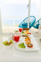 Una mesa de comedor con diversos alimentos y bebidas, vista a la playa a través de amplios ventanales, y sillas azules.