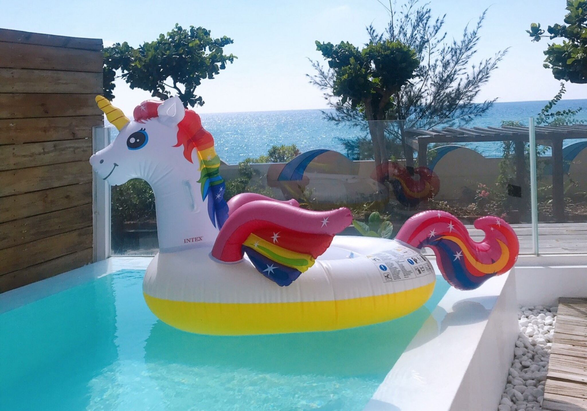 Een opblaasbaar eenhoorn drijvend in een zwembad met een houten terras en uitzicht op zee.