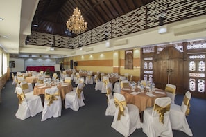 A banquet hall with round tables set for events, white chairs with gold sashes, a wooden partition with intricate designs, and a chandelier.
