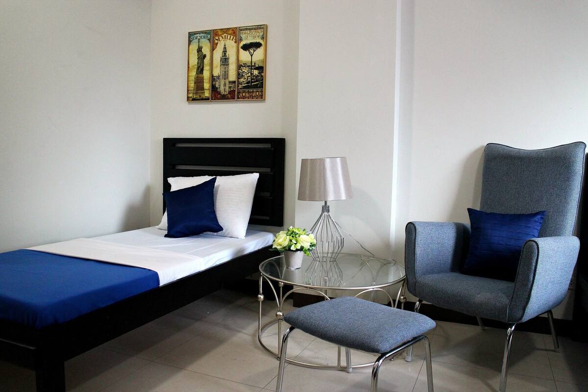 A room with a bed, a chair, a small table with a lamp, and a wall decoration.