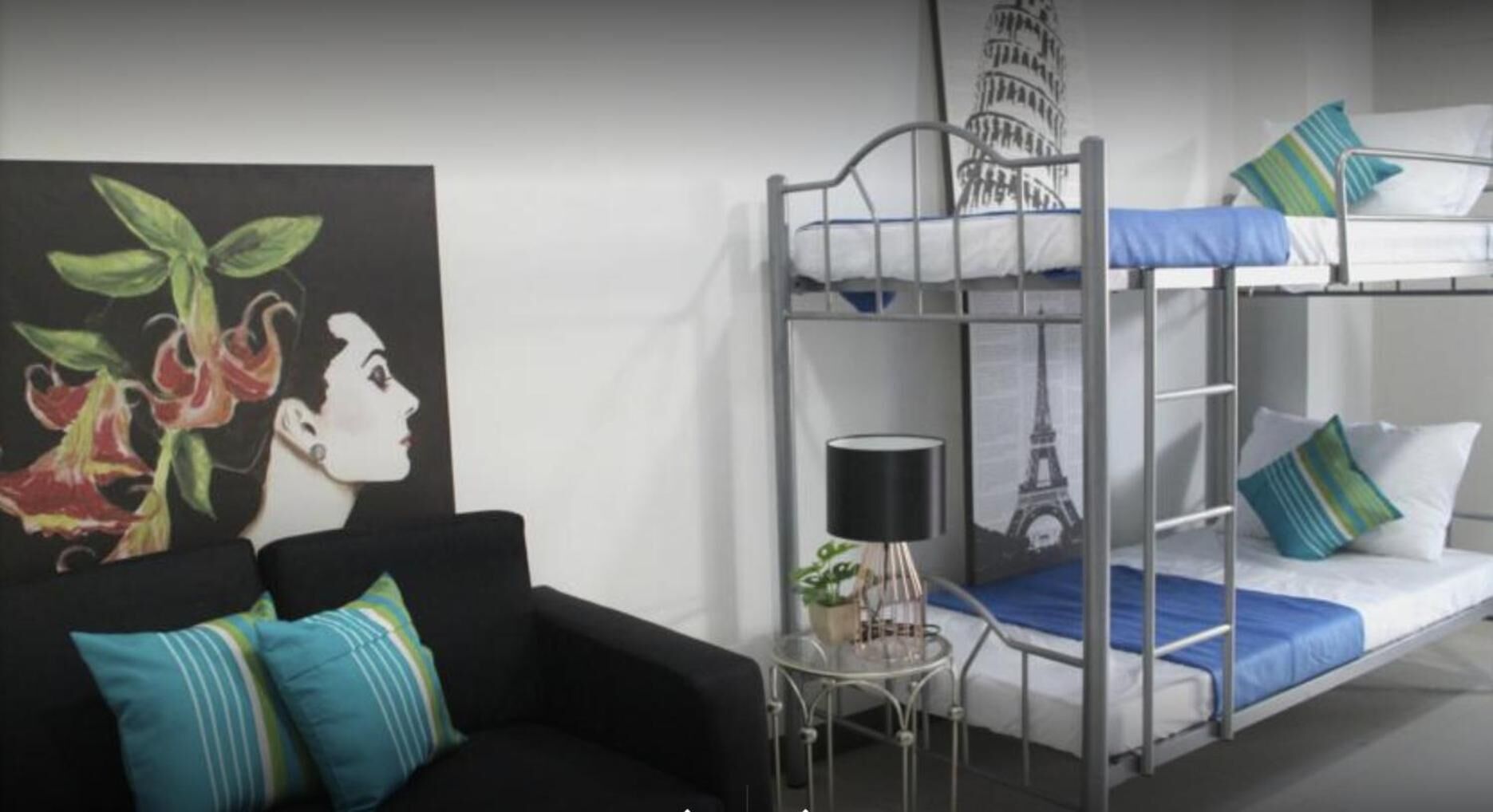 A bunk bed with a metal frame, a small table with a lamp, a black couch with striped pillows, and a painting of flowers and a woman's face.