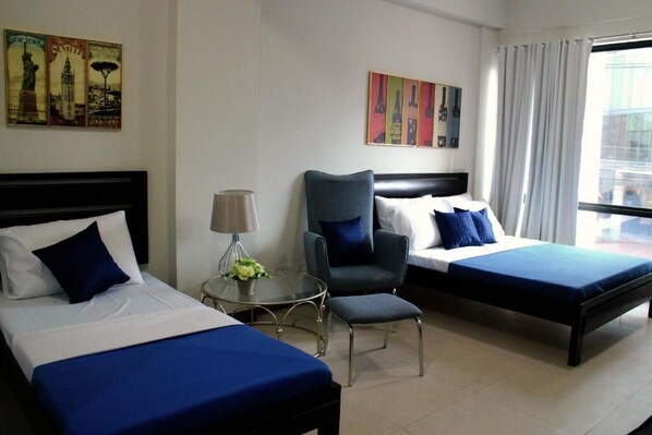 Una habitación de hotel con dos camas, una silla, una mesa con lámpara y obras de arte en las paredes.