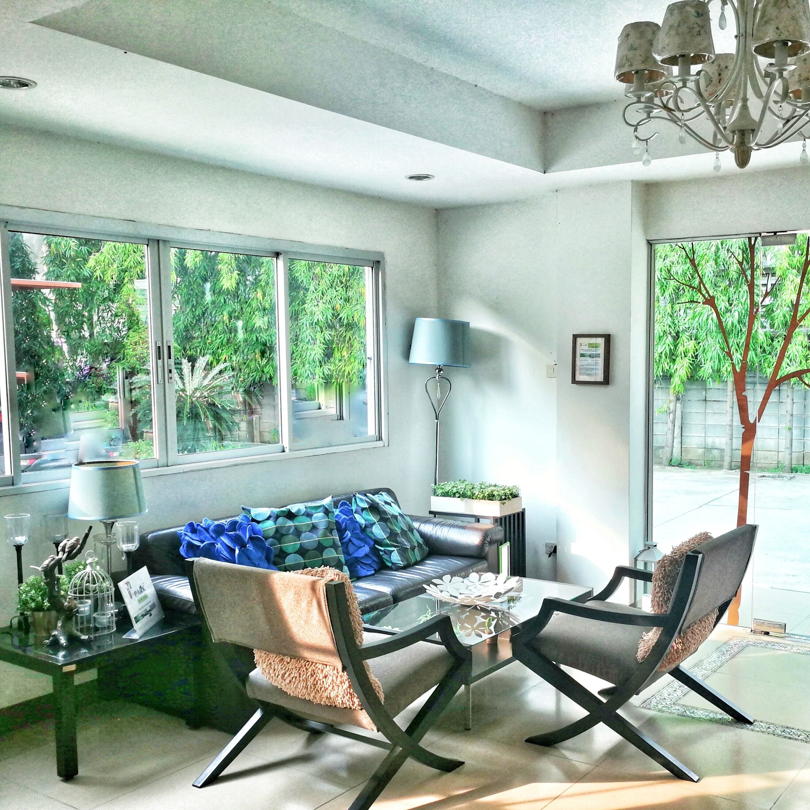 A modern living room with a sofa, two armchairs, a coffee table, and a chandelier. Large windows offer views of greenery outside.
