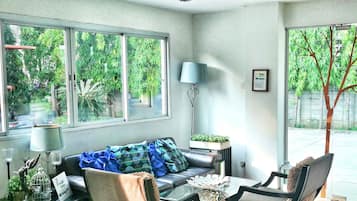 A modern living room with a sofa, two armchairs, a coffee table, and a chandelier. Large windows offer views of greenery outside.