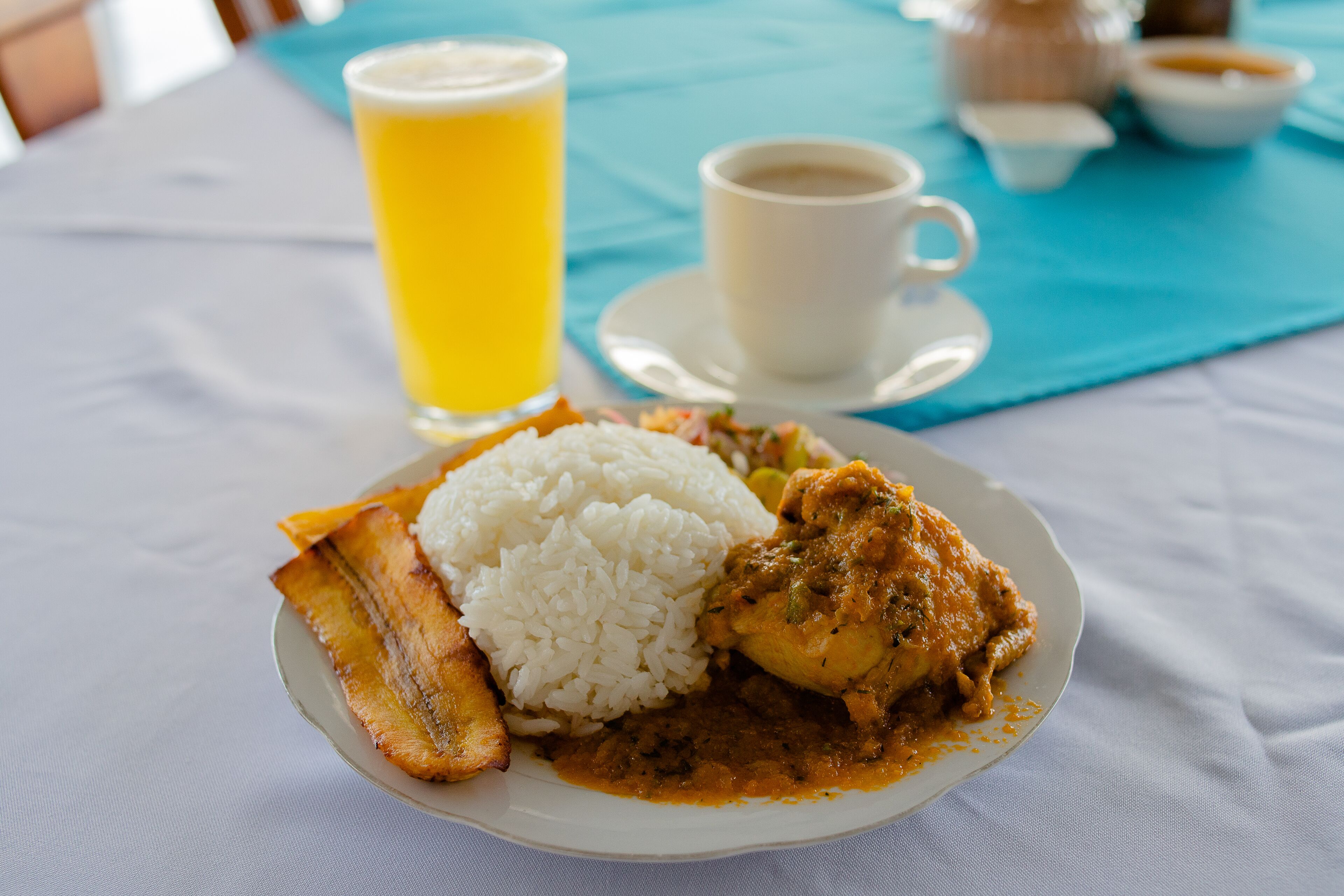 Un plato de comida con arroz, plátanos y un curry, un vaso de jugo de naranja y una taza de café sobre una mesa.