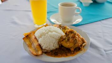 Un plato de comida con arroz, plátanos y un curry, un vaso de jugo de naranja y una taza de café sobre una mesa.