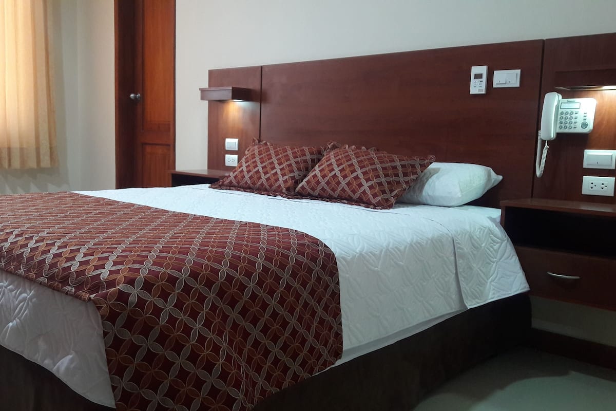 Una habitación de hotel con una cama, una mesita de noche con teléfono y un termostato fijado a la pared.