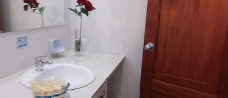 Une salle de bain avec un lavabo blanc, un miroir, une porte en bois et un vase rempli de fleurs rouges.