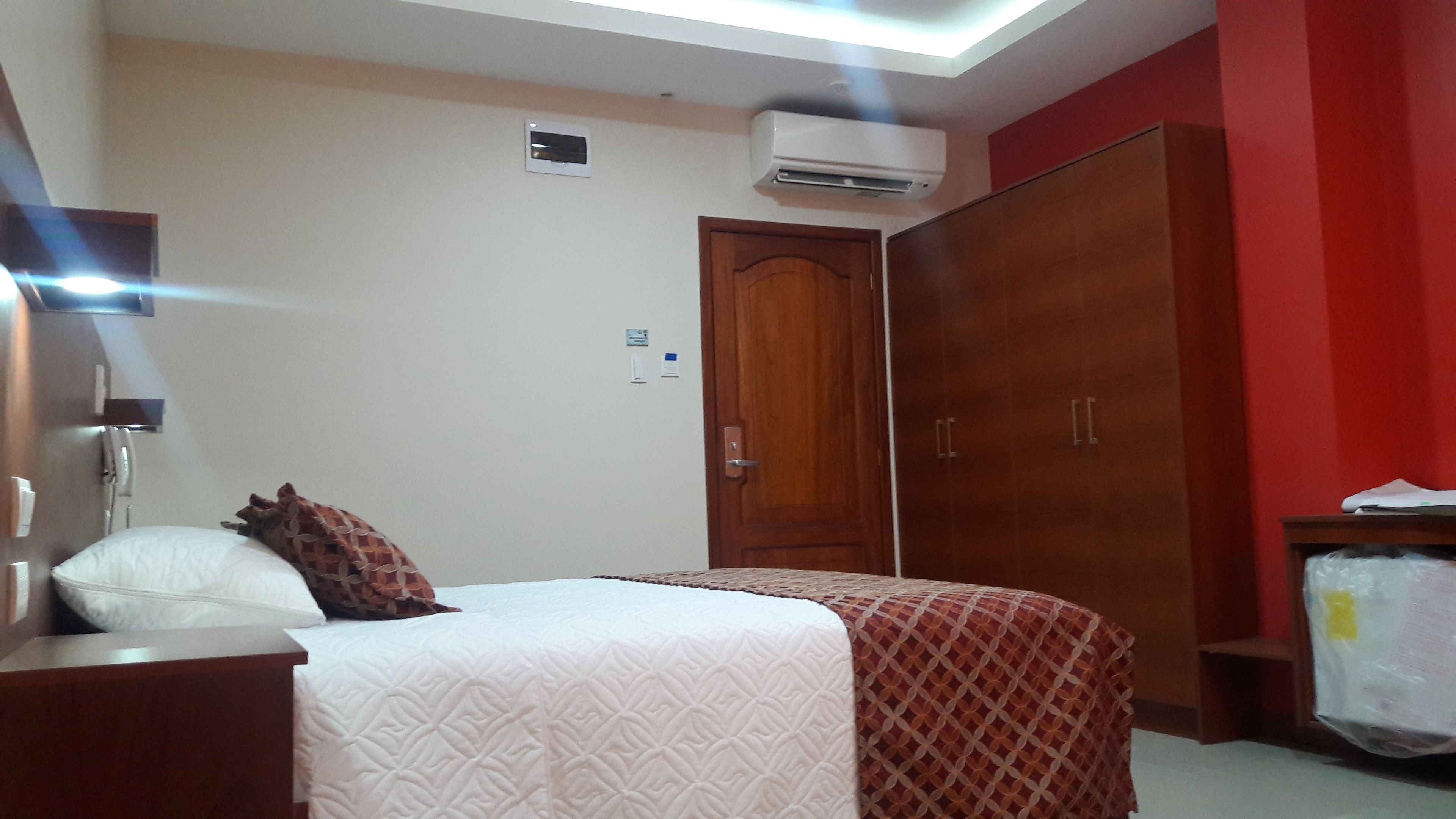A hotel room with a bed, a wardrobe, an air conditioning unit, and a bedside table.