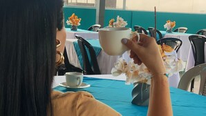 A person seated at a table with a white tablecloth, holding a white cup. The table is set with a blue table runner and has a vase with flowers.
