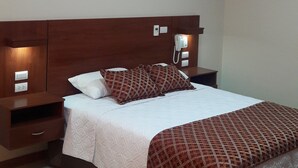 A hotel room with a wooden headboard, a bed with patterned bedding, a bedside table with a phone, and a wall socket.