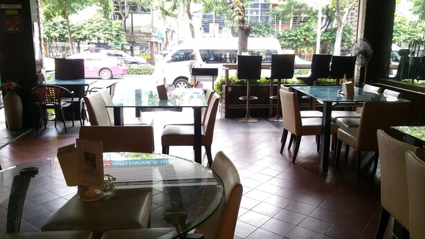 An outdoor cafe with glass tables and chairs, a tiled floor, and a view of the street with cars and trees. - Relax Time Silom - Hostel (Bangkok)