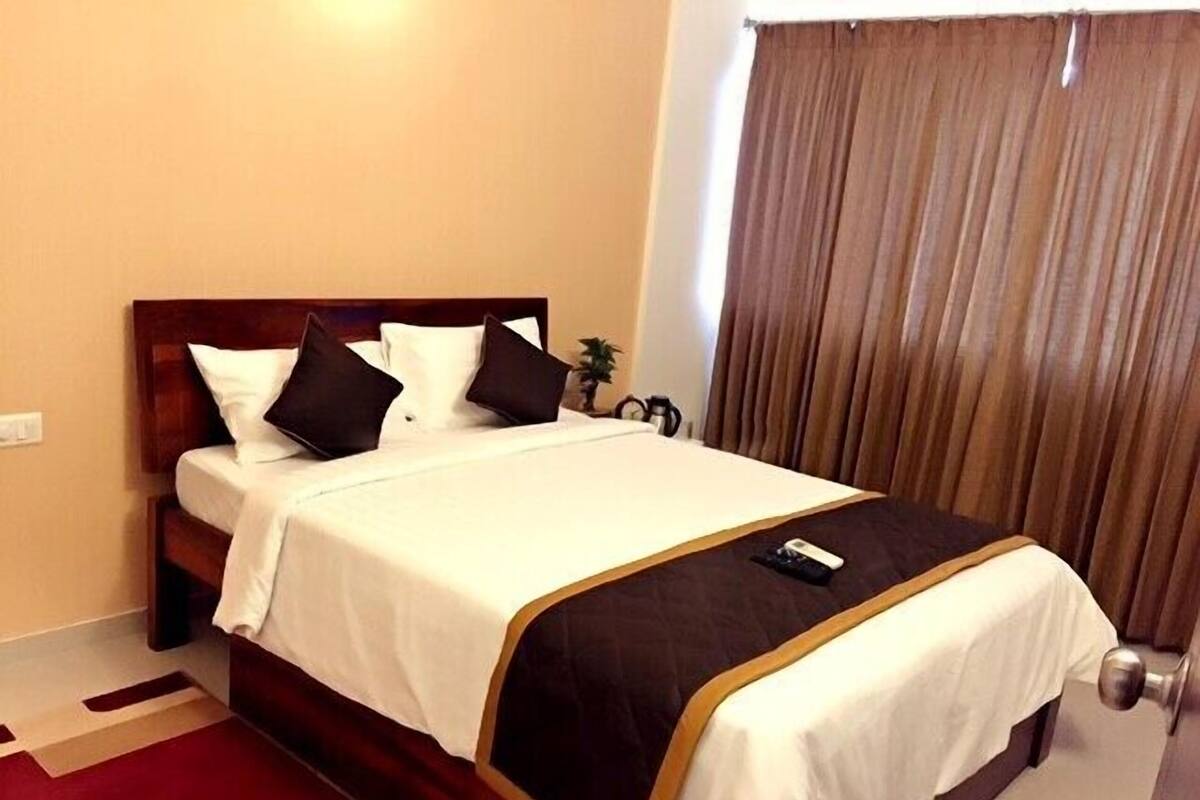 Una cama bien hecha con ropa de cama blanca y marrón, un control remoto sobre la cama y una alfombra estampada en tonos rojo y beige.