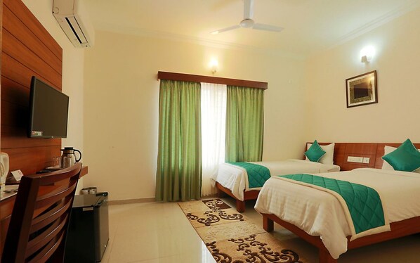 A hotel room with two beds, a TV, a desk with a coffee maker, and a window with green curtains.