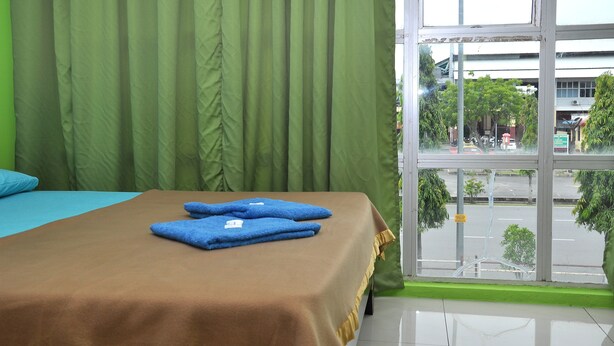Une chambre d’hôtel avec un lit, des rideaux verts et une vue sur la rue, visible à travers une grande fenêtre.