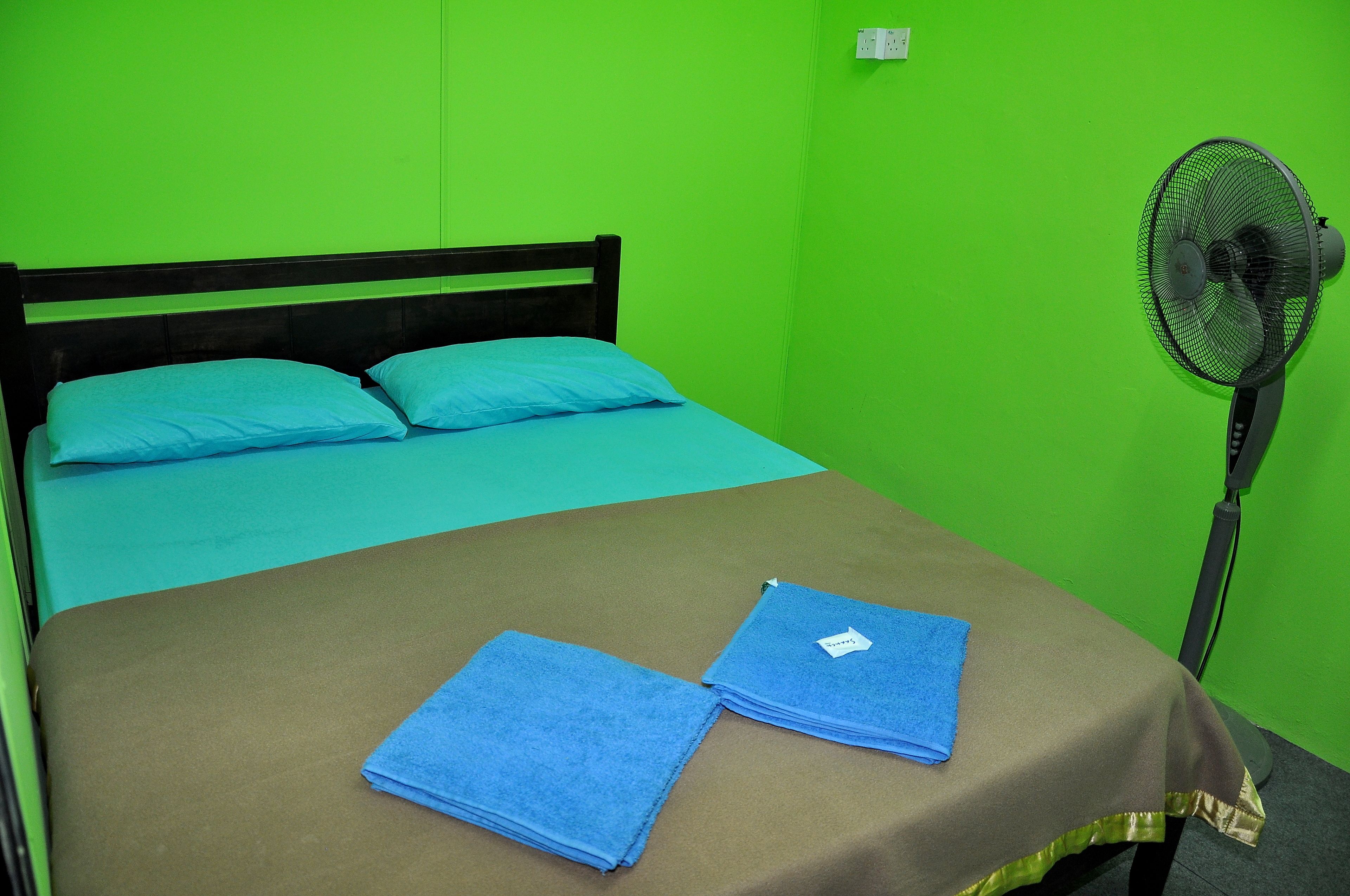 A single bed with a blue mattress and pillows, a brown bedspread, and two folded blue towels on the bed. A standing fan is positioned to the right of the bed.