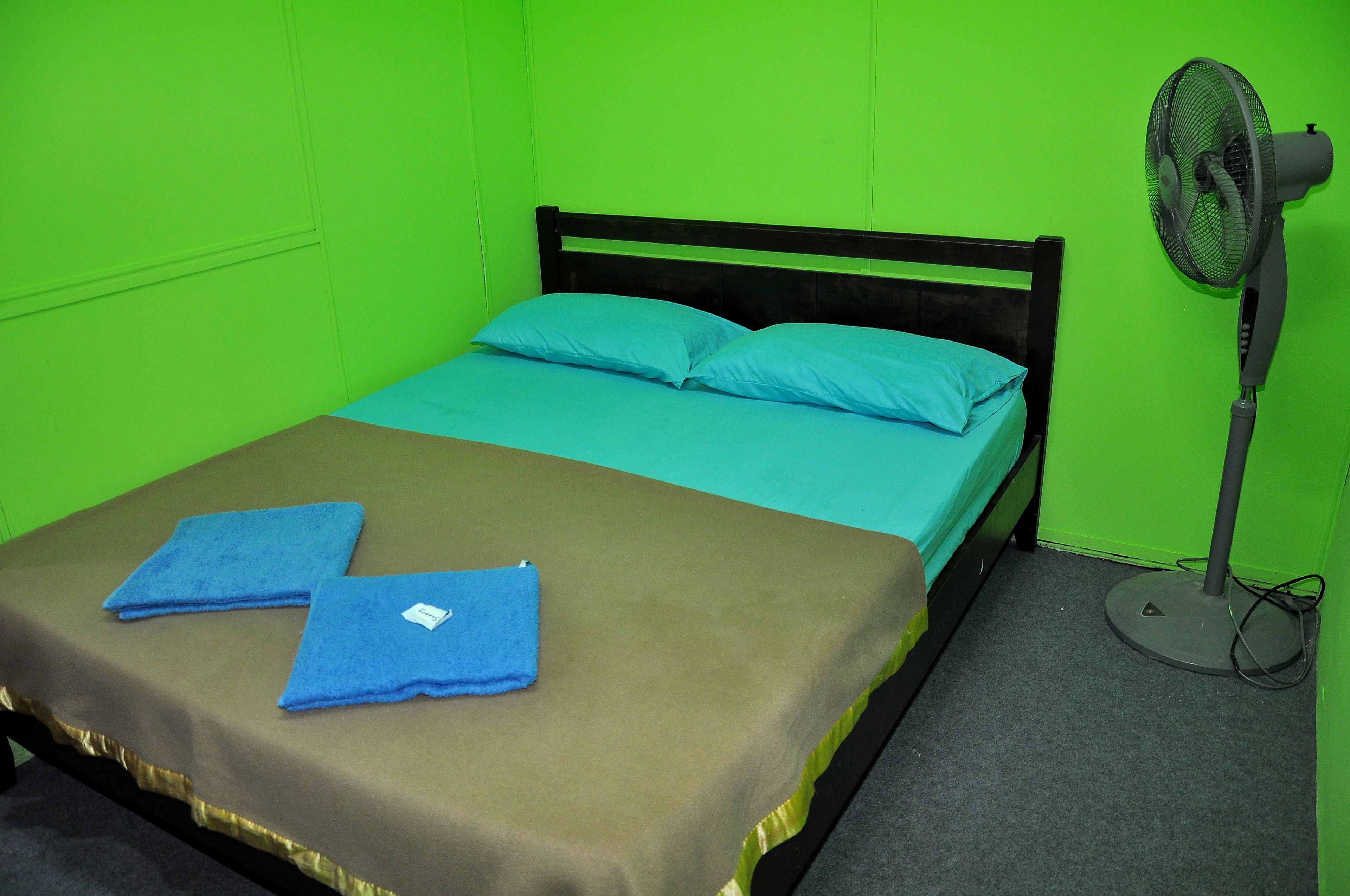 A single bed with a brown bedspread and two blue pillows, a standing fan, and a pair of blue towels on the bed.