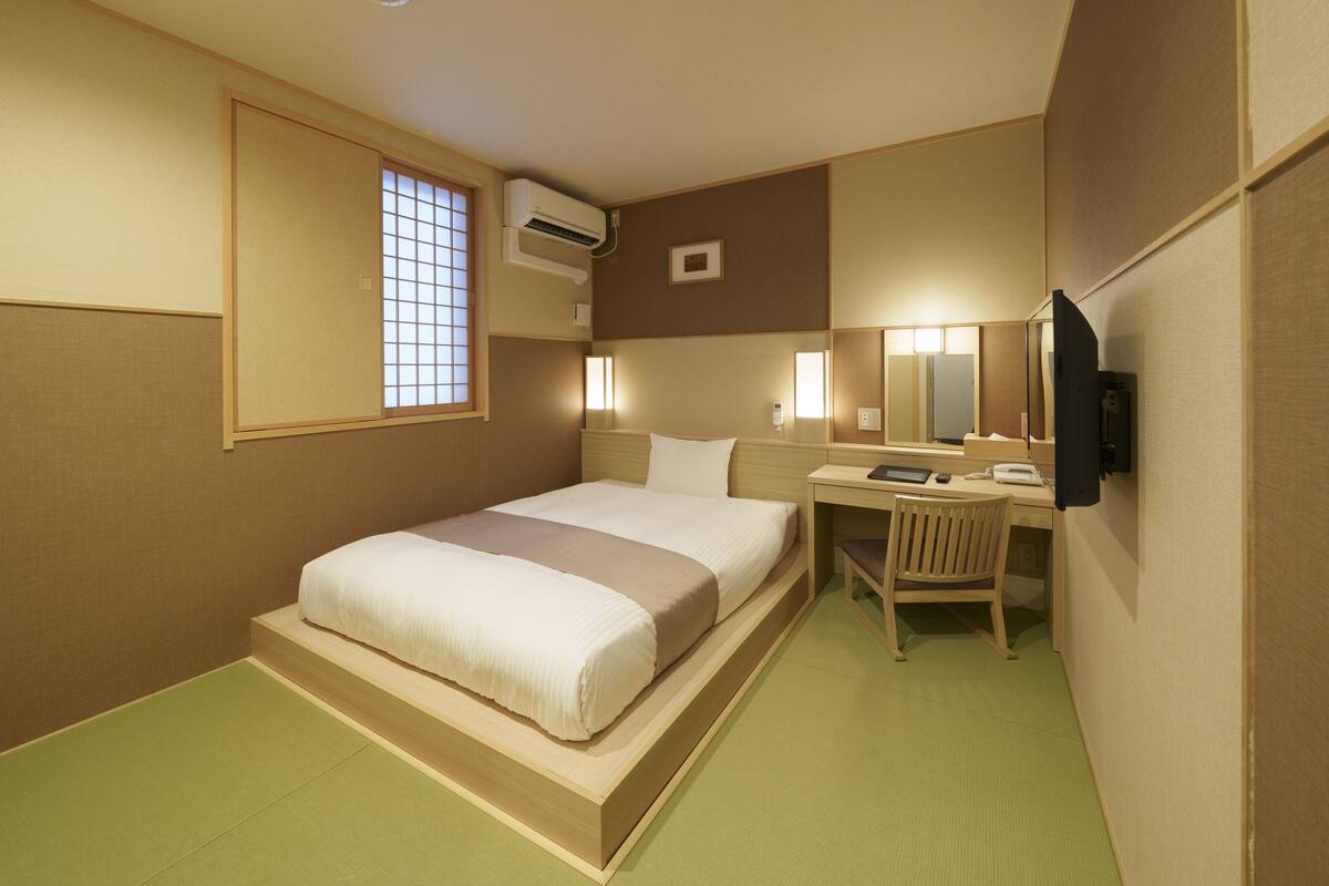 A hotel room with a bed, a desk, a chair, a television, and an air conditioning unit.